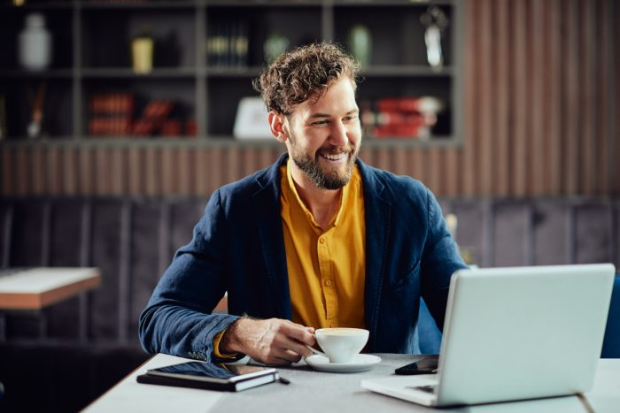 Bearded smiling Caucasian freelancer sitting in cafe, drinking fresh coffee and using lapotp.